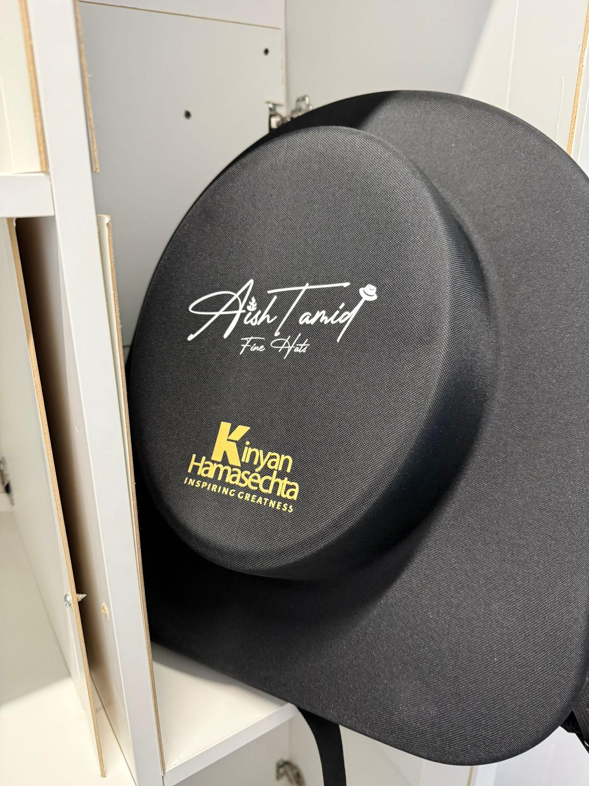 Aish Tamid hat travel bag with the gold Aish Tamid Fine Hats emblem and a customer's Kinyan Hamasechta logo printed beneath it